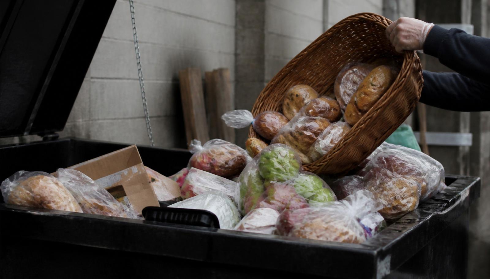 Baskets of perfectly good sealed bread and packaged food being tossed into a restaurant dumpster.